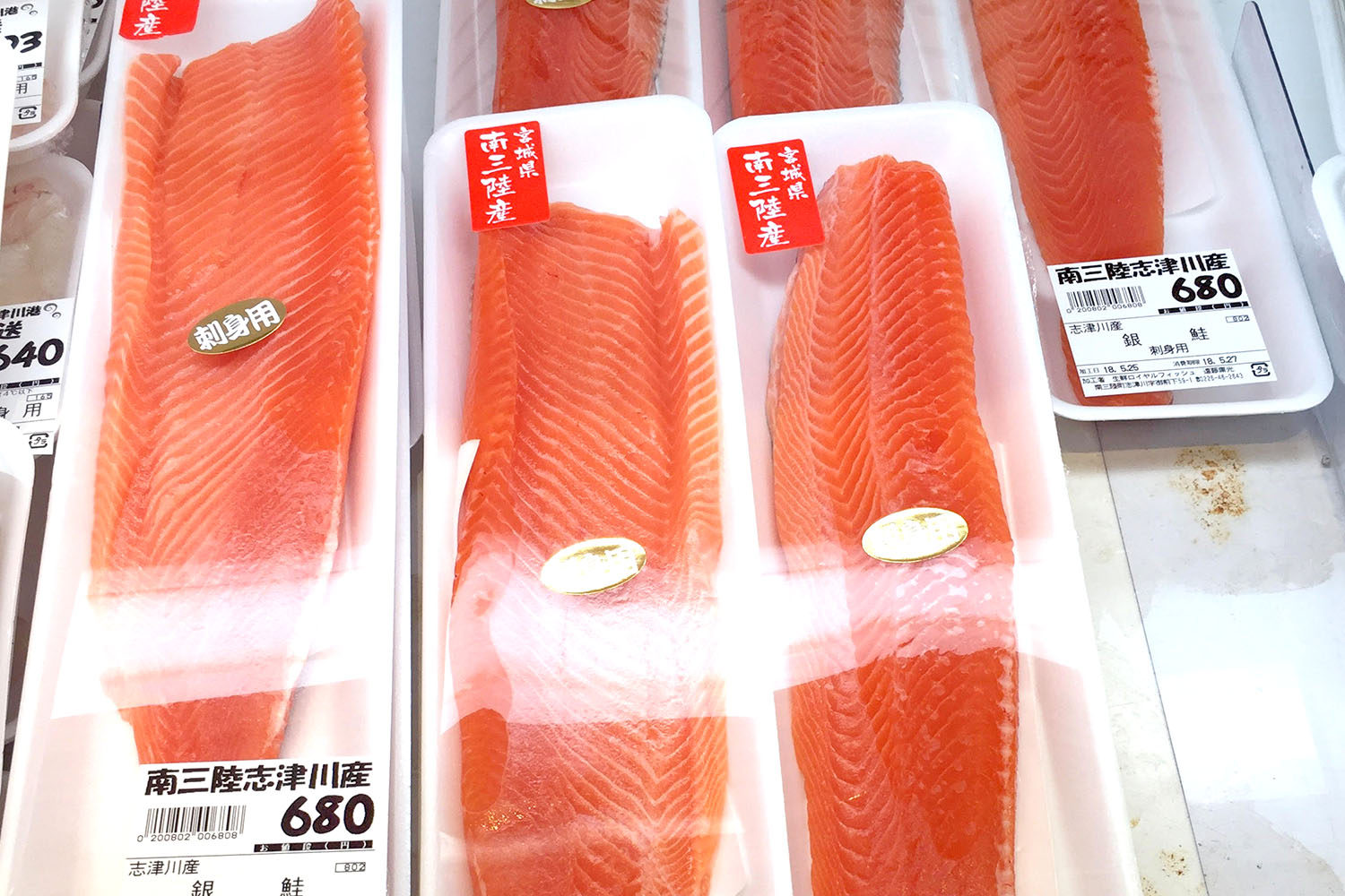 スーパーの鮮魚売り場に、価格が「680円」と表示されているプラスチックトレイに入った赤みがかった新鮮なサーモンの切り身が並んでいる写真