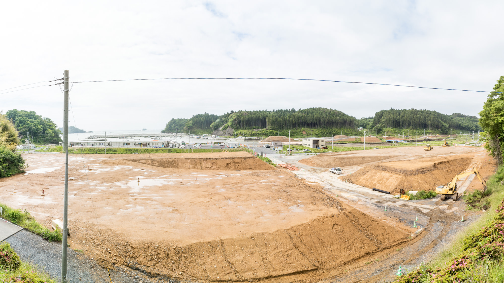 明るい曇り空の中定点カメラによる風景で広大な造成地が広がっており、測量用の目印（赤い旗や杭）がいくつか見られ、これから造成区画が細かく決められるような写真