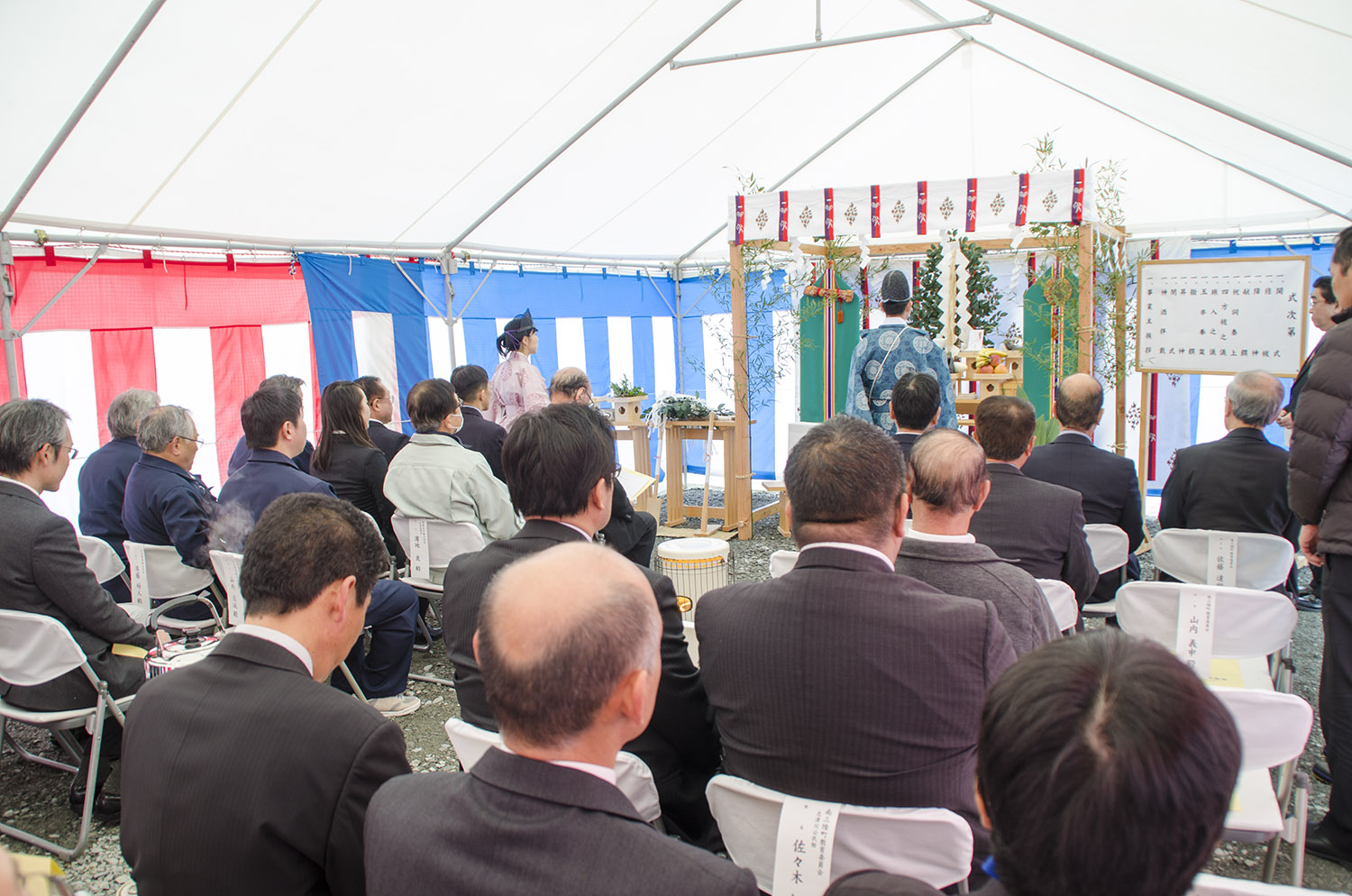屋外の白いテント内で椅子に座った多くの男性や女性が祭壇の方向を向いて座っており、祭壇には供え物が飾られていて、正装した神官が祭礼を執り行っている様子が写っている安全祈願祭の写真