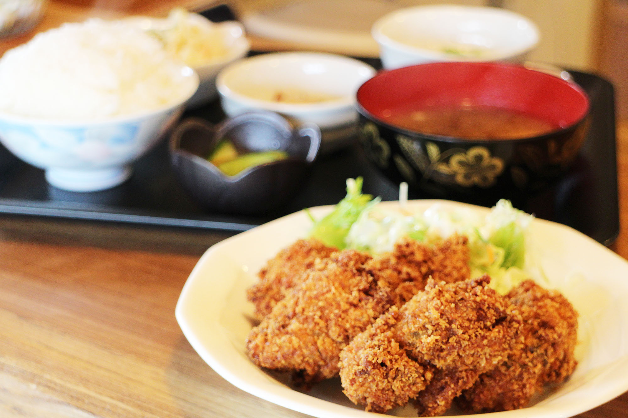 ご飯、味噌汁、小鉢が乗った黒いお盆と、手前に大きなカキフライと千切りキャベツが盛られた白い皿が置かれている写真