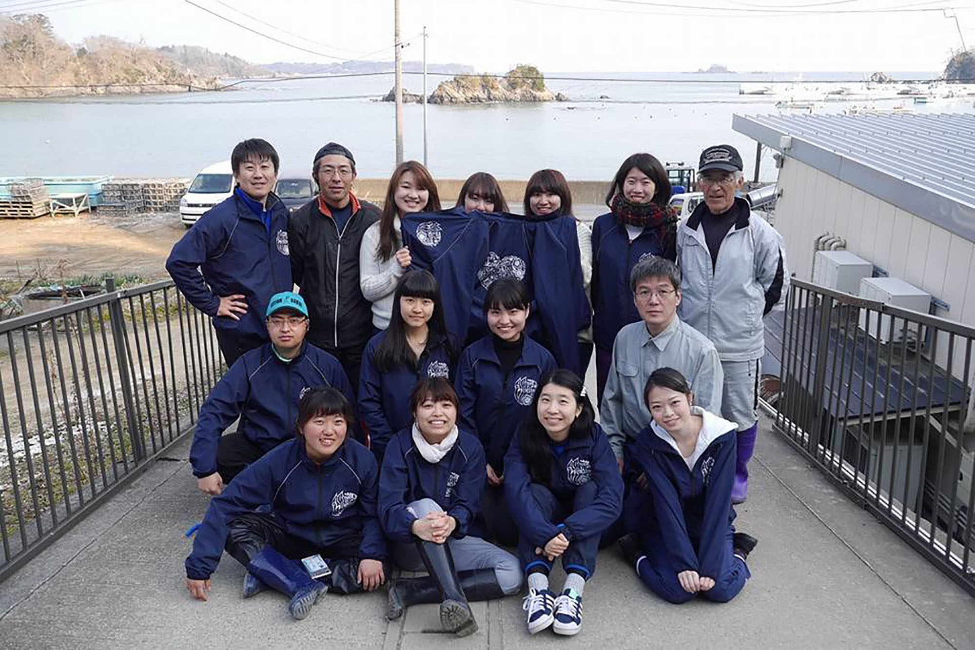 海辺の風景を背景に、前方の8人が座り、後方に7人が立って笑顔で集合写真を撮っている写真