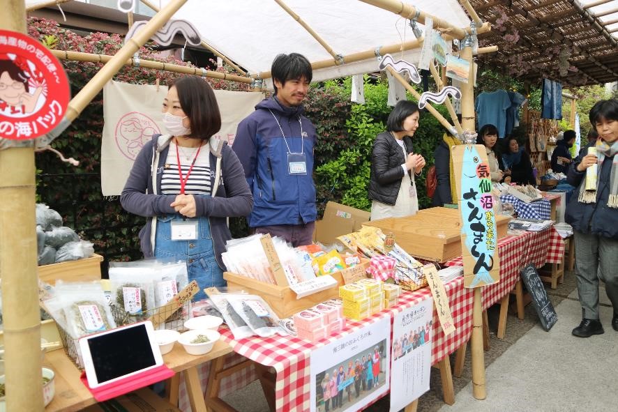 赤いチェック柄のテーブルクロスの上に加工食品や小物が並び、名札をつけた出店者たちが笑顔で来場者を迎えている販売ブースの写真