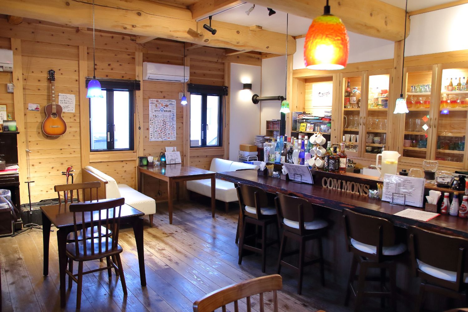 木の温もりを感じる店内に、白いソファと木製テーブル、ギターが壁にかかっていて、カウンターの後ろには酒瓶が並び、色とりどりのペンダントライトが天井から吊り下がっているカフェの全景の写真