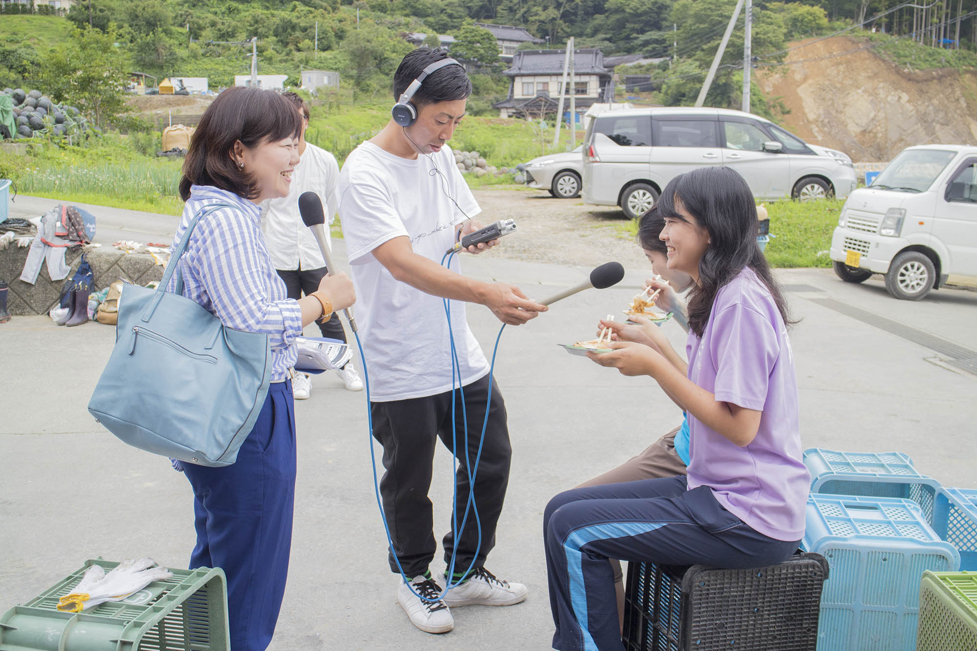 屋外で空のコンテナに座り食事をしている恵泉女学園高等学校OGの2名に、マイクを持ったリポーターの女性とヘッドホンをして2人にマイクを向けている男性が取材をしている様子の写真