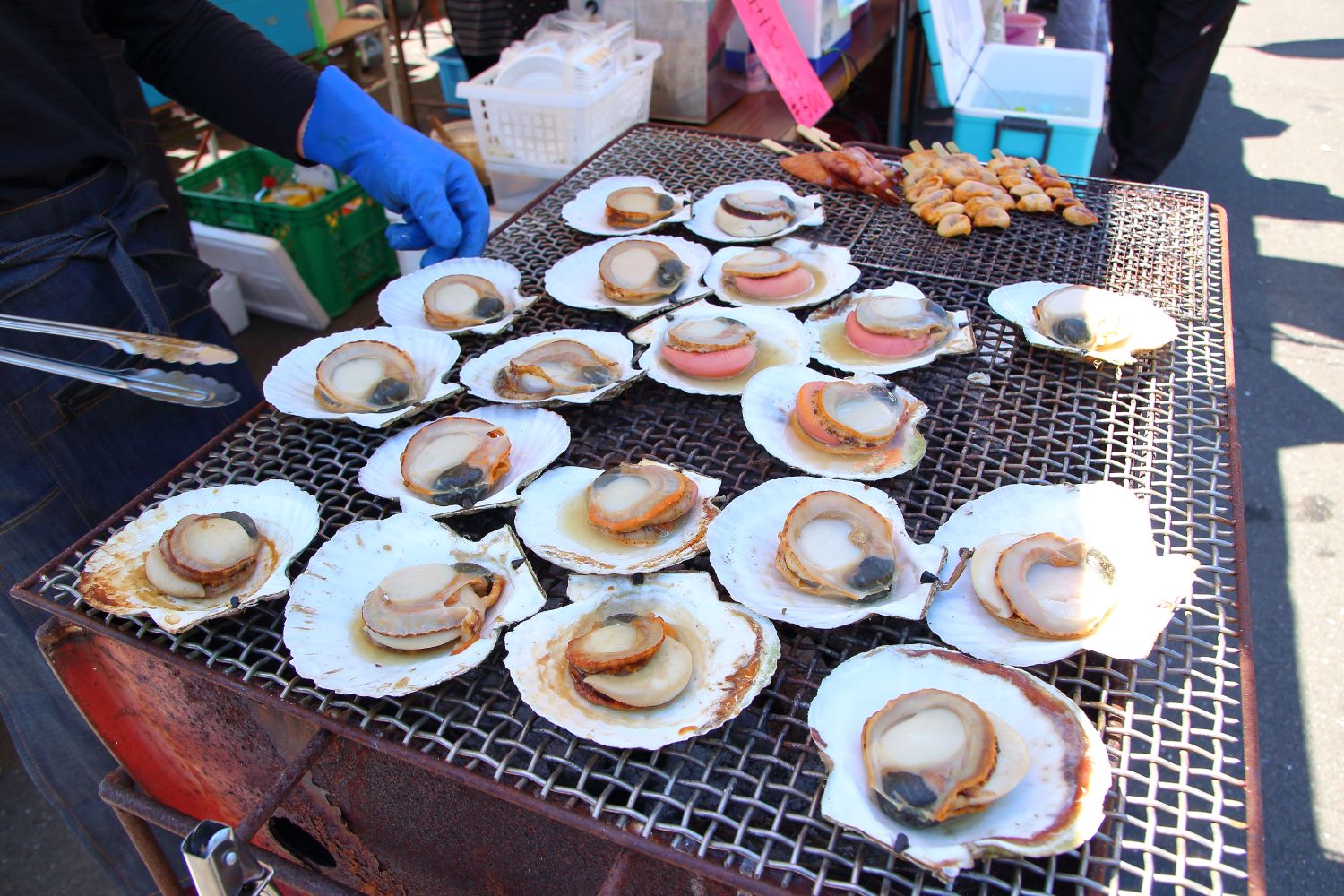 鉄網の上に貝殻ごと乗せたホタテが並び、焼かれながらじゅうじゅう音を立てており、手袋をした人がトングで扱っている屋台の調理中の写真