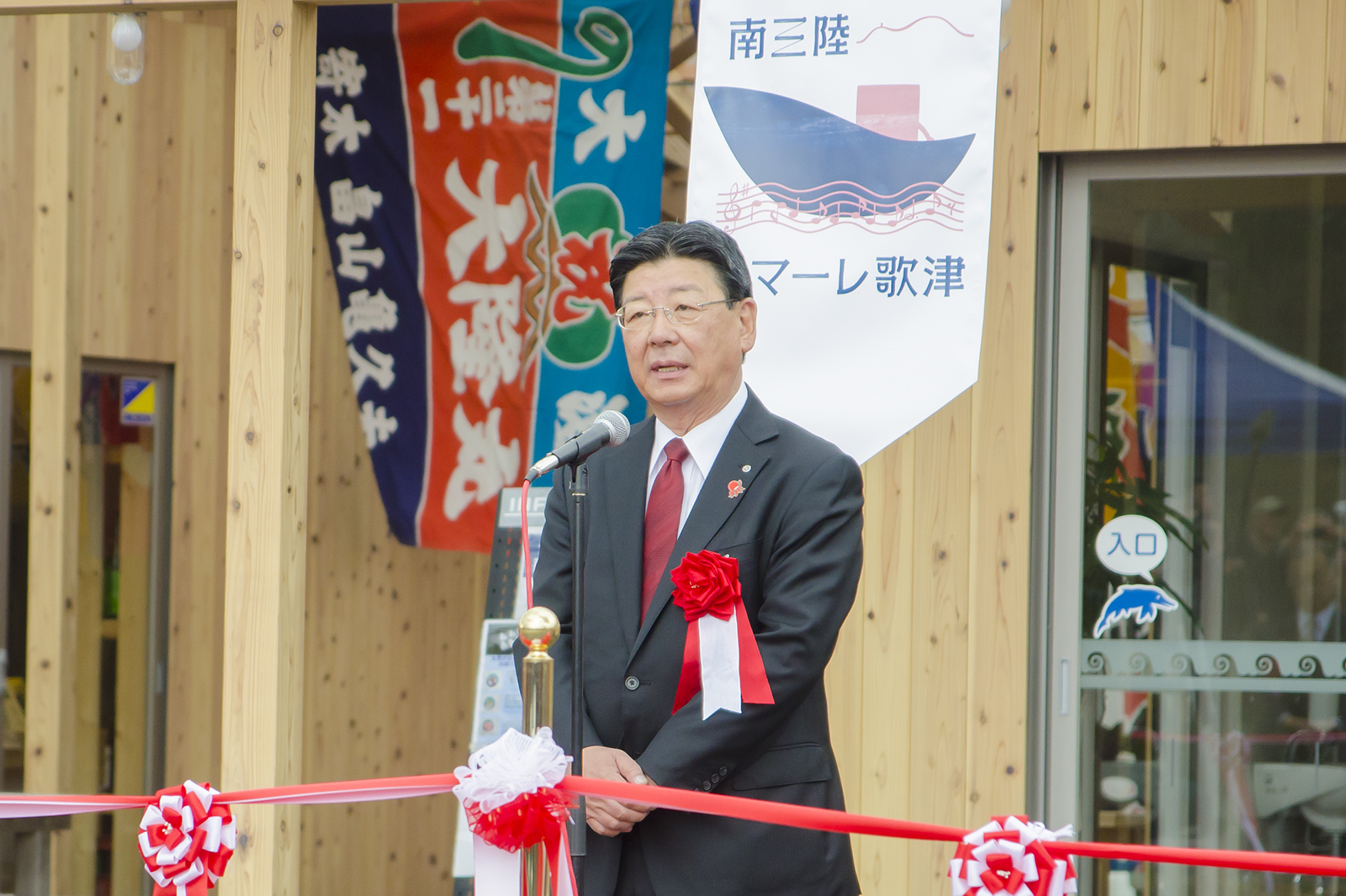 真新しい木造の建物の前に立ち、紅白花飾りがついたテープの後ろに置かれたマイクの前で話をしている佐藤町長の写真