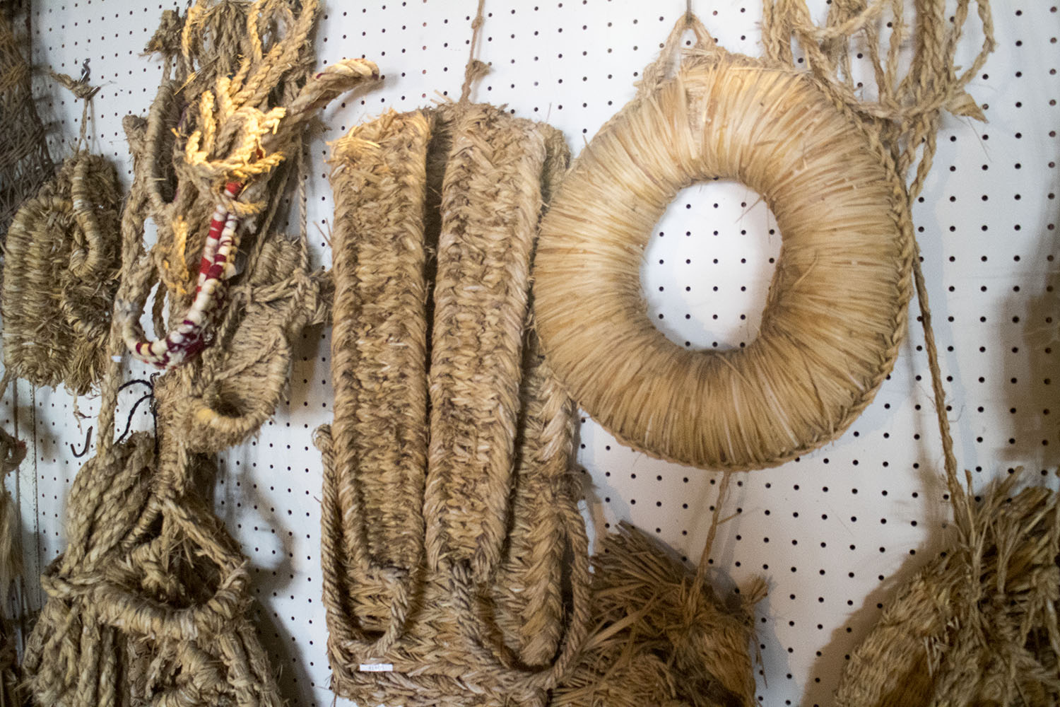 輪の形をしたしめ飾りや、厚みのあるぞうりのような形、縄状に編まれたものなど、さまざまな形状のわら細工が壁に並んで展示されている様子の写真