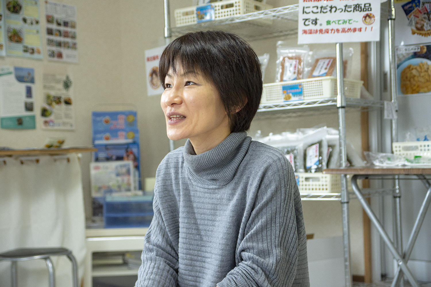 棚に並べられた食品やポスターの前で、グレーのタートルネックを着た女性が笑顔で話している様子を斜め前から撮影した写真