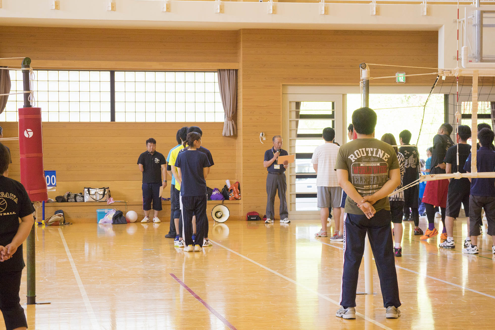 体育館の中で、複数の参加者が一列に並んで立ち、前方でマイクを持った男性が話をし、参加者たちが話を聞いている様子の写真