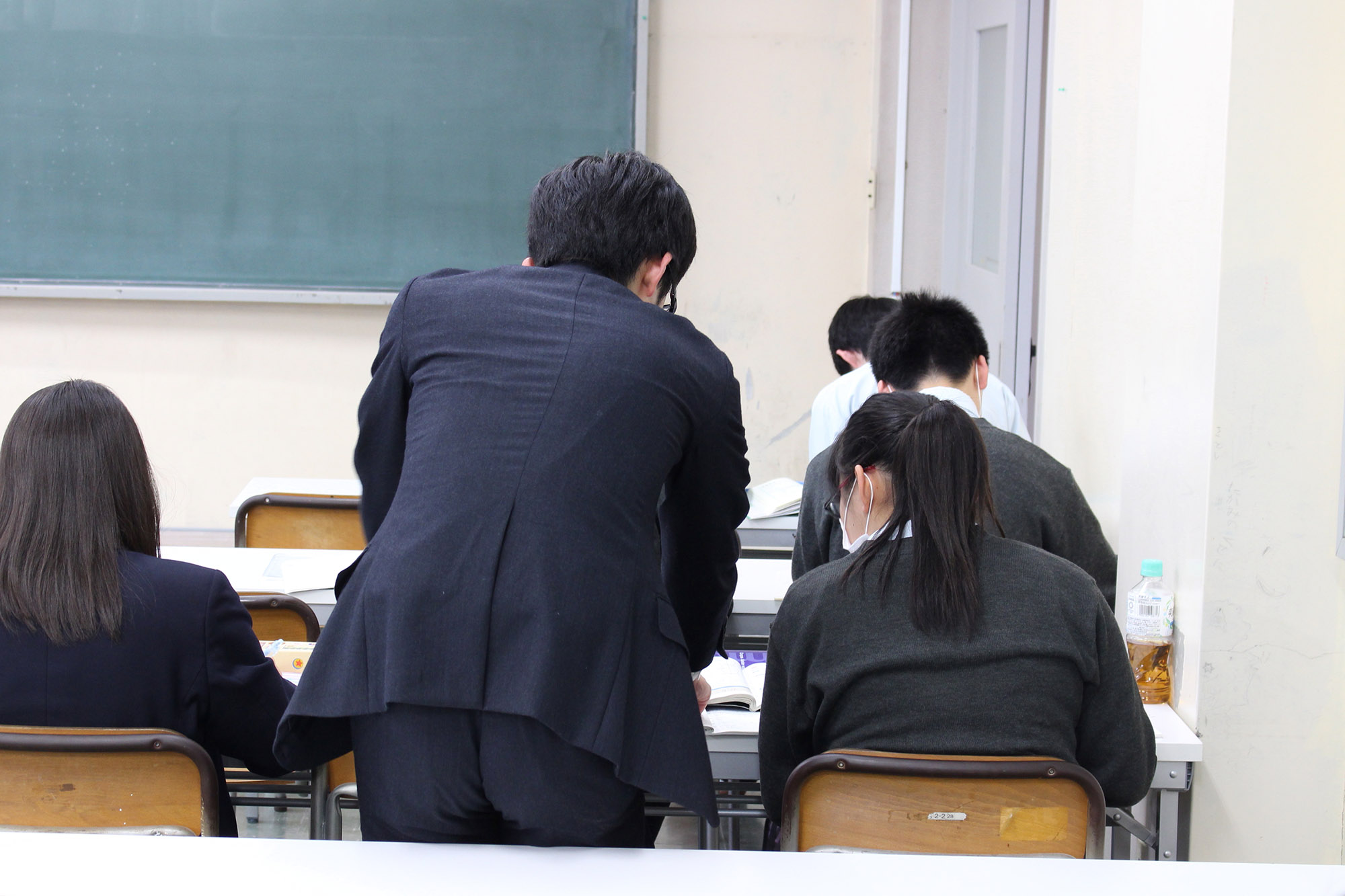 教室内に並べられた机に向かって学習している女子生徒の横にスーツを着た男性が立ち、女子生徒に学習支援をしている様子の写真