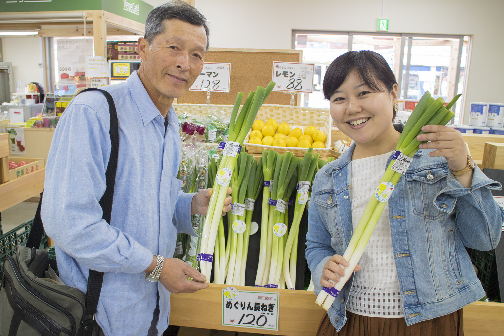 野菜売り場のネギが陳列された棚の前で長ネギを持った阿部さんと女性リポーターが笑顔で写っている写真