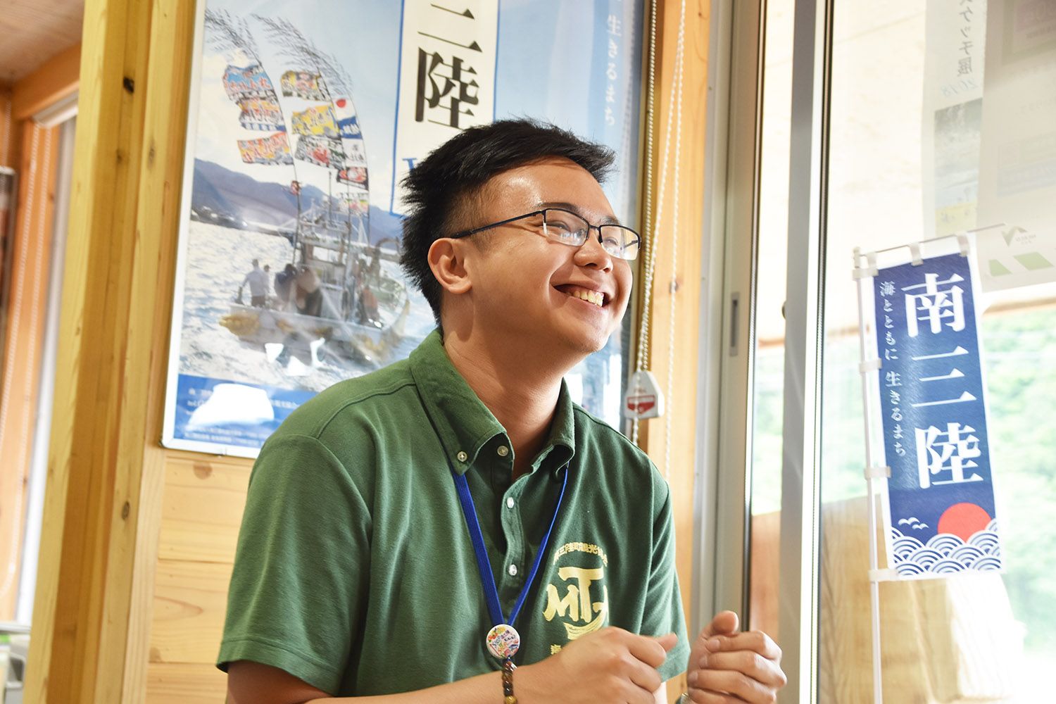 背後には漁船の写真ポスターと「南三陸」と書かれた青い旗が飾られている木造の観光施設の室内で、緑のポロシャツを着た陳さんが笑顔で話をしている写真