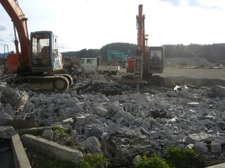 がれきや建材の破片が散らばる屋外の作業現場で、2台の重機が作業を行っている様子の写真