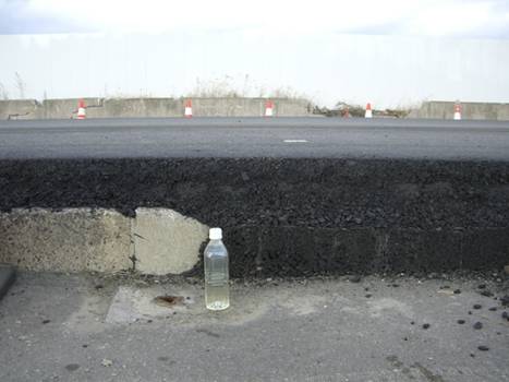 道路の一部が約1メートル程度地盤沈下しており、低くなった路面にペットボトルが立てられて沈下の深さを示し、奥には高い路面の縁に沿って交通コーンが並べられている、地盤沈下の状況を記録した写真