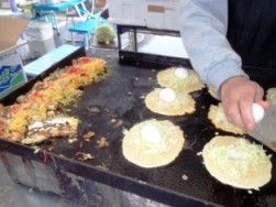 屋台の鉄板焼きで、生地の上にキャベツと卵がのった料理が調理されている様子を写した写真