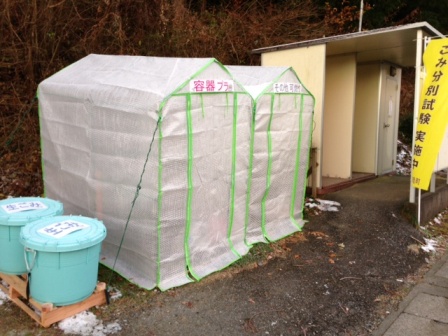 白いカバーのついたな屋根付きの緑色フレームの回収ボックスに『容器プラ』と『その他プラ』の表示があり、左側には青いごみ箱、右側には『ごみ分別試験実施中』と書かれた黄色い垂れ幕が掲げられている、ごみ分別回収のモデル事業の写真