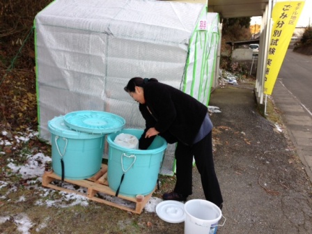 雪が残る屋外で、水色の大型ごみ箱にごみを入れている女性の写真