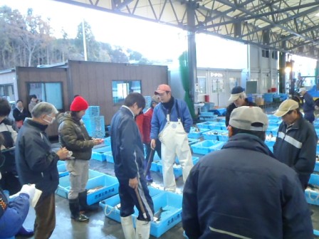 魚の入った青い容器の周りに人々が集まり、何人かがメモ帳や紙を持っている様子が写っており、魚の競りが行われている様子の写真