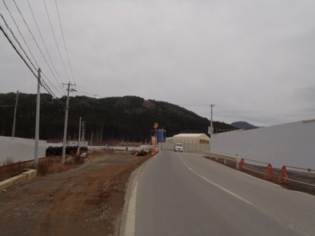 舗装道路の両側に白い仮囲いの塀が続き、その先に災害廃棄物処理施設が見える写真