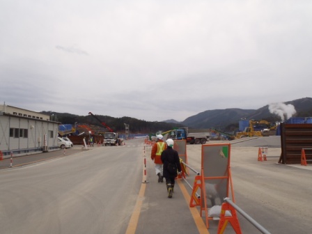 舗装道路をヘルメットと作業着を着た二人の作業員が歩いており、左には仮設の建物や車両、右奥には大きな機械や重機が稼働している様子の写真