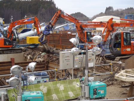 オレンジ色の重機が3台並び、アームで木材などのがれきをつかんで選別しており、手前には投光器や発電機などが設置されている写真