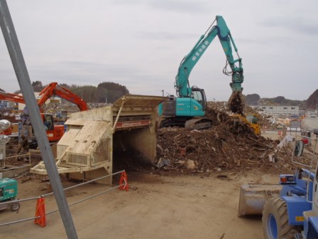 青緑色の大きなショベルカーが、大きな山のように積まれたがれきの上で作業をしている写真