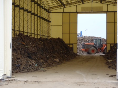 白い大型テント内の左右に木くずなどの廃材が高く積まれ、奥の開口部からは外に停まっているオレンジ色の重機と廃棄物の山が見える写真。