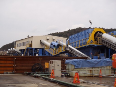 白い壁の建物から使途に伸びたベルトコンベヤーに青と黄色の配色の機械が接続されている処理施設の写真