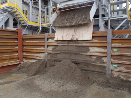 上の階から土砂が滑り落ちるシュートの先に仕切り壁で囲まれたエリアがあり、そこに洗浄済みの土が山になって積もっていて、背景には金属製の構造物や黄色い手すりが見える写真