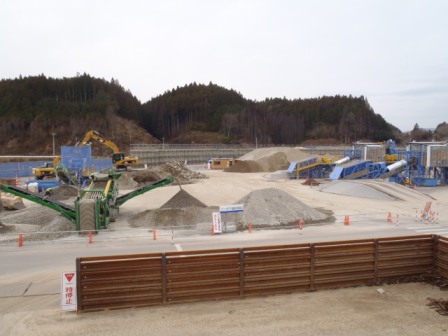 手前に鉄製の仕切りがあり、その向こうには広い作業ヤードが広がり、複数のショベルカーやブルドーザーが稼働していて、大小さまざまな砂利や土の山が点在している様子の写真