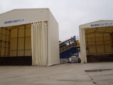 白いシートで囲まれた二つの大型テントが建ち、左のテントには「建設副産物（汚泥）テント」、右のテントには「建設副産物テント」と書かれ、それぞれの中には黒っぽい土や資材が保管されており、奥には黄色い手すりがついた青いコンベア設備が見えている写真