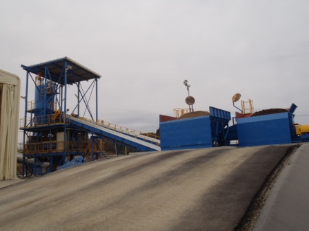 土が入った青色の大きなコンテナの手前にゆるやかなスロープ状のコンクリート舗装があり、左側には屋根付きのタワー状構造とコンベアベルトが見える写真