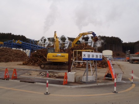 木破砕施設の看板が設置された場所に木材が積み上げられ、黄色いショベルカーが作業を行っている様子の写真