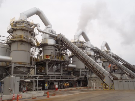 金属製の筒状構造を持つ焼却設備が屋外に3基並んで設置され、それぞれの排気筒から煙が立ち上り、斜めに延びるベルトコンベヤーが手前に設置され、地面には作業員や安全コーンが見える写真