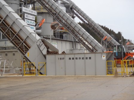 「1号炉 廃棄物供給装置」と書かれた灰色の箱型設備に、ベルトコンベヤーを通じて木くずのようなものが運ばれ、奥には同じ装置が2基とショベルカーが見える写真