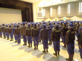 大勢の制服を着た消防団と交通指導隊の人々が整然と並んで立っており、敬礼をしている後ろ姿を写した写真