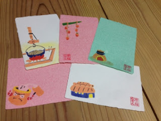 白、ピンク、緑の手漉きハガキに囲炉裏や吊るし柿、藁ぶき屋根の家などがデザインされ赤い印鑑が押されているものが5枚並べて置かれている写真