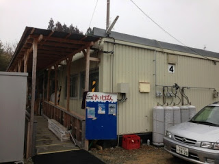 白いプレハブ造りの建物で、建物の正面には木の柱とトタン屋根でできた簡易なひさし付き通路があり、入口付近に青い掲示板とプロパンガスボンベが設置された仮設住宅の写真