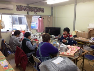 テーブルを囲んで4人の人々が机の上に置かれたミシンや布を使って作業したり談笑したりしているプレハブの室内の写真