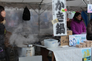 「かき鍋」と書かれた看板を下げた白いテントの中で大きな鍋から湯気が立ち上っている様子の写真
