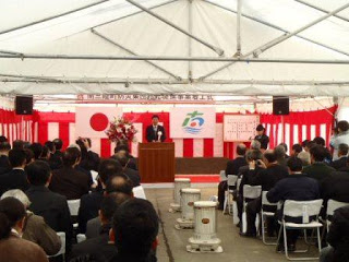 白いテントの中に紅白幕が張られ、日の丸と町旗が掲げられた壇上の演壇に立った男性の話を椅子に座った多くの出席者が聞いている様子の写真