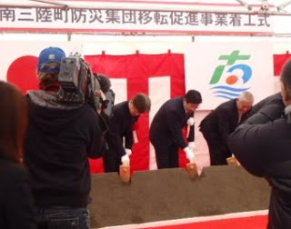 「南三陸町防災集団移転促進事業着工式」と書かれた横断幕と町旗が掲げられた紅白幕の前でスーツ姿の男性数名が鍬入れをしており、その様子をカメラマンが撮影している写真