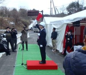 白いテントの前で、数台のカメラで撮影する中、赤い台の上に立った白いヘルメットをかぶった男性が赤い旗を高く掲げて号令をかけている様子の写真