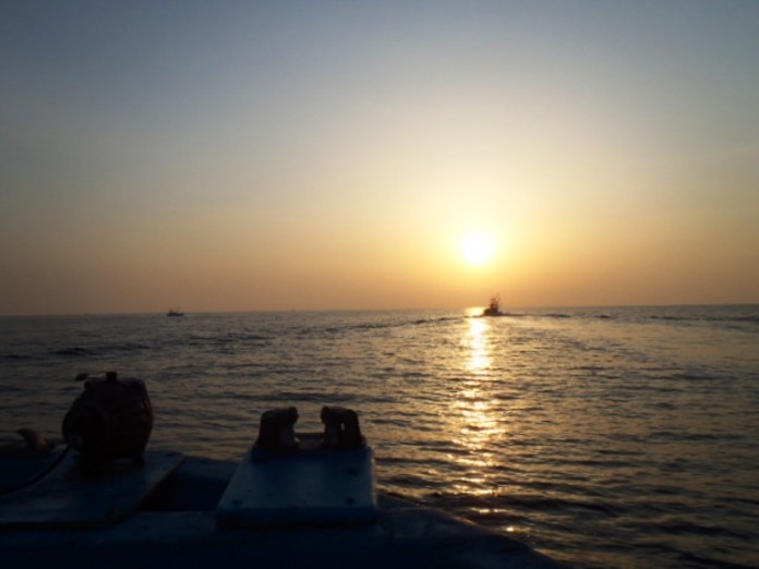 海の上に太陽の光がまっすぐ金色の帯のように伸び、水平線の近くに浮かんでいる一隻の漁船のシルエットが黒く浮かび上がっている写真
