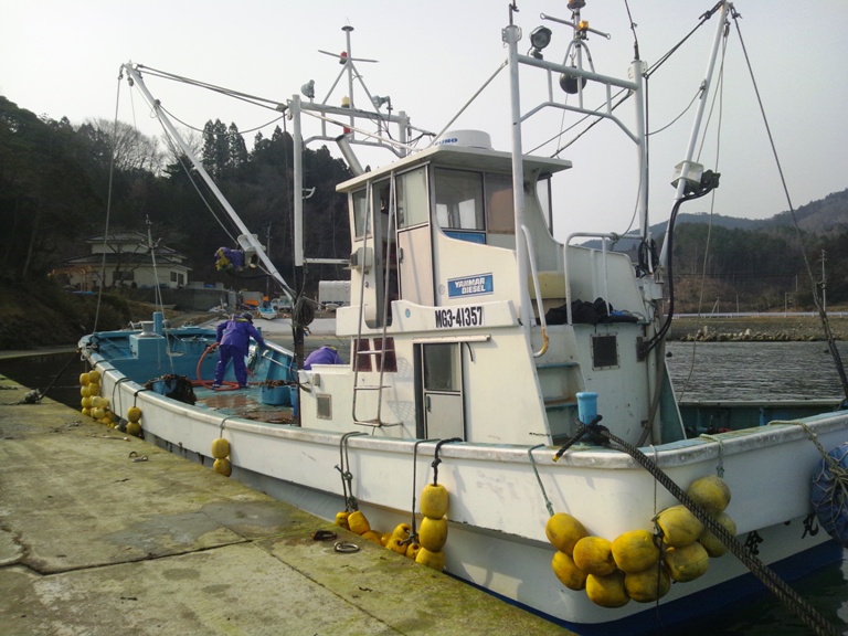 側面に黄色いブイがずらりと並んだ白い漁船の甲板で、紫の作業着を着た漁師がデッキを清掃している写真