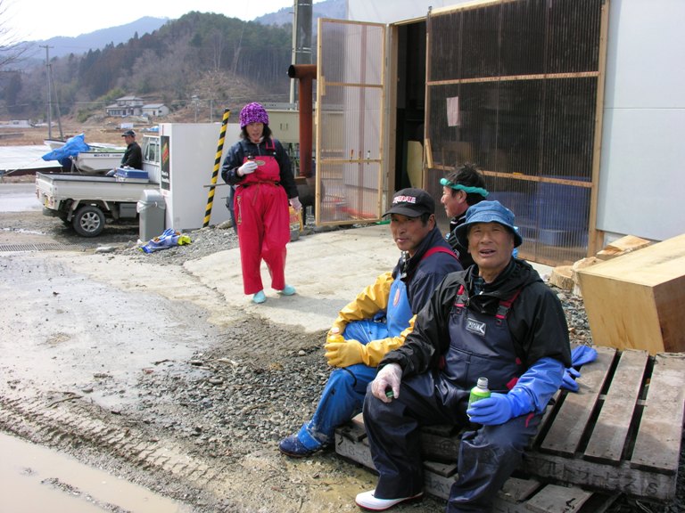 作業着を着たわかめ漁の作業員たちが施設の外で休憩をとり、飲み物を片手に笑顔で談笑している様子の写真