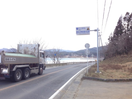 右側に山と草地、左側に海が見える緩やかなカーブの国道45号線を大型ダンプカーが走っており、道路の右側には「ここから過去の津波浸水区間」と書かれた標識が掲げられている写真