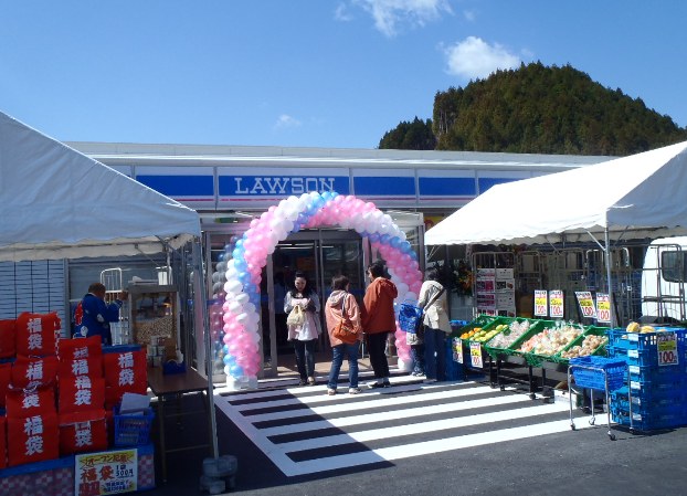 青空の下、山の近くに新たにオープンしたローソンの店舗前にピンクと白の風船アーチが飾られ、店の前に設置された2つのテントの下には野菜や福袋が並び、買い物客が出入りしている様子の写真
