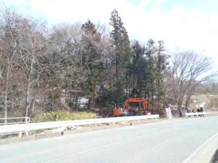 背の高い杉の木などが生い茂る林が道路沿いにあり、その前でオレンジ色のショベルカーによる作業が行われていて、周囲には数人の作業員が作業をしている様子の写真