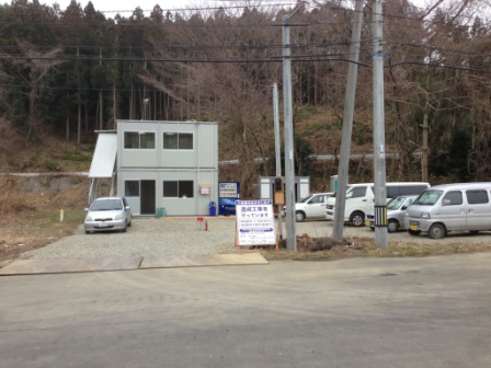 森を背後に建てられた簡易的なプレハブの事務所があり、その前に複数の車が駐車している造成工事の場所を写した写真